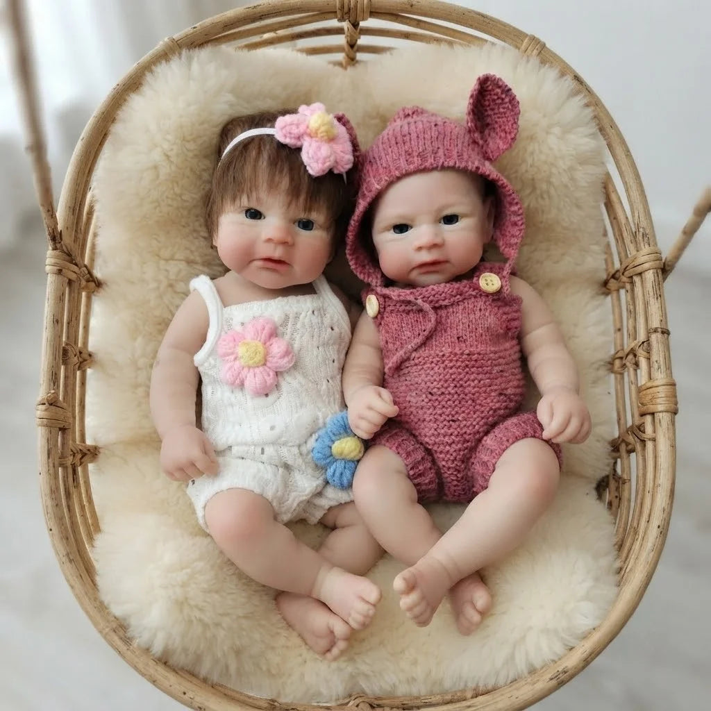 A realistic soft silicone reborn doll with pink hair and ears is seated in a woven basket lined with fluffy cream fabric.