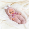 A pink fluffy reborn baby doll wearing a matching pink hooded outfit is resting on white fur.