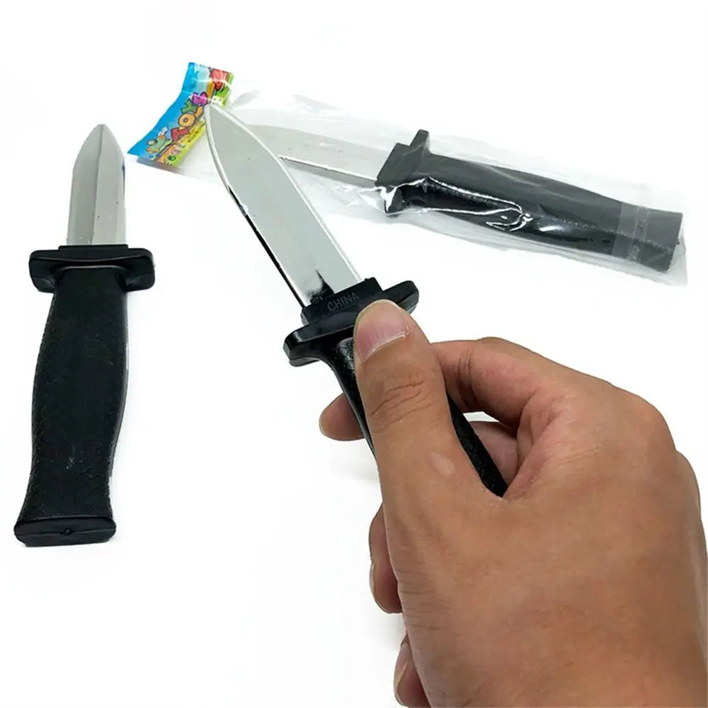 A hand holds a silver retractable trick dagger prop knife with a black handle and blade, packaged in plastic.