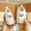 A pair of white lace-trimmed baby girl moccasin shoes with bows being held by a person's hands.