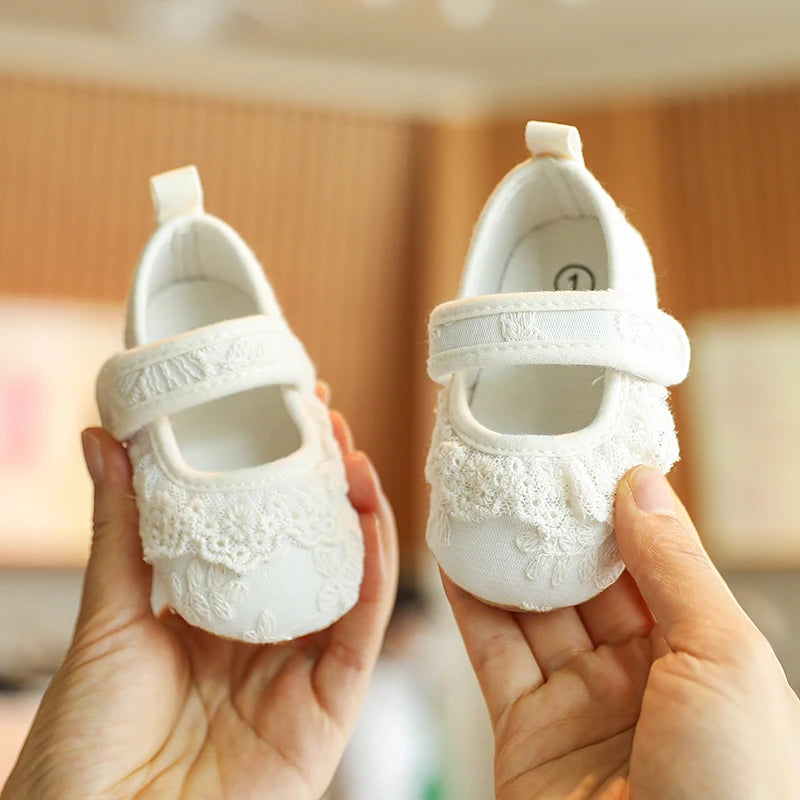 A pair of white lace-trimmed baby girl moccasin shoes with bows being held by a person's hands.