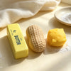 A yellow butter stick, a beige food-shaped squishy, and some white powder are arranged on a light surface.