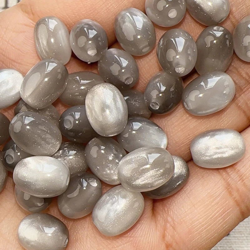A hand holds a pack of ten iridescent, silvery-gray resin star-like cat's eye beads for jewelry making.