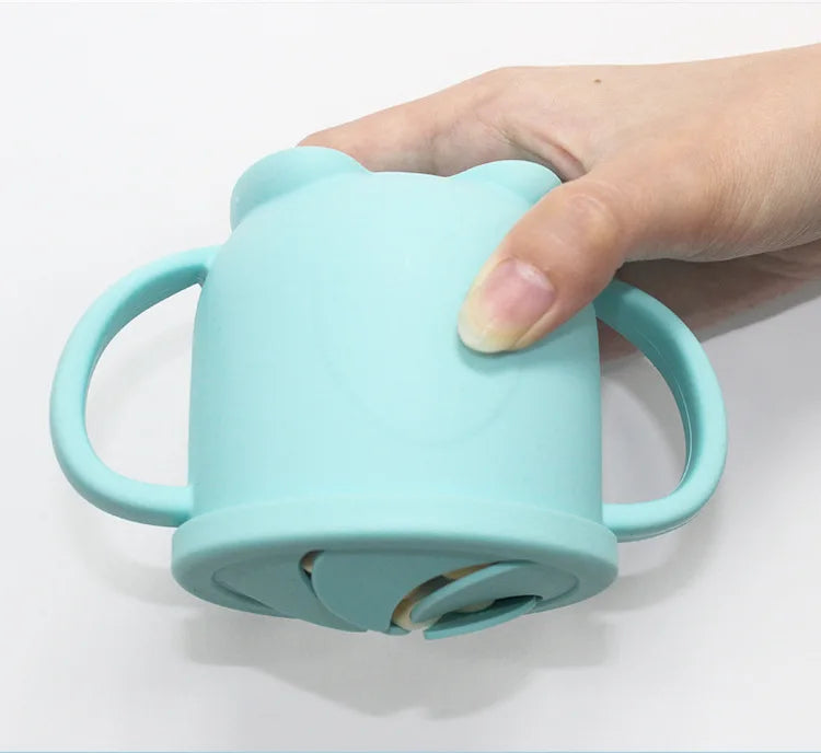 A hand is holding a light blue silicone snack cup with a lid for babies and toddlers.