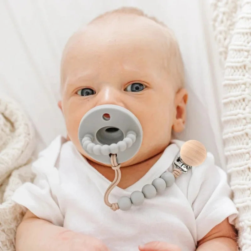 Baby trägt eine Silikon-Trinkfläschchen-Klemme an der Brust, weißer Hintergrund und gestricktes Material sichtbar.