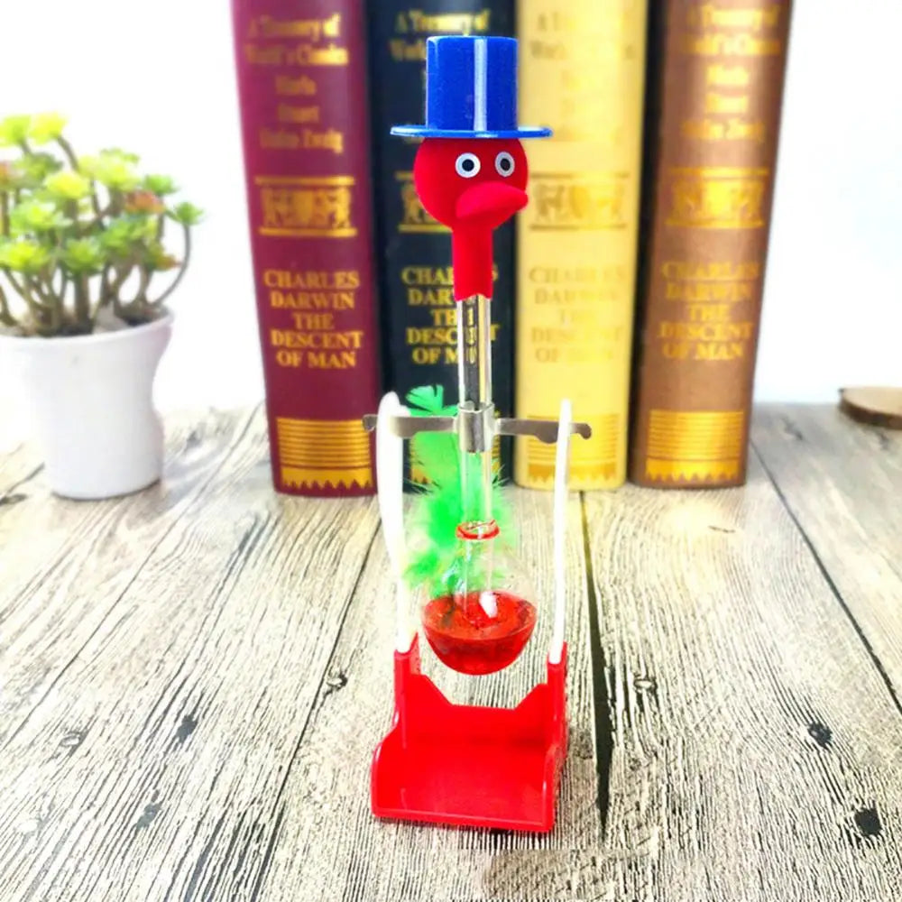A red and blue drinking bird toy with a long neck and green feather is displayed in front of old books on a wooden surface.