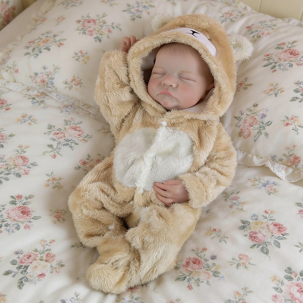A realistic vinyl reborn baby doll wearing a tan bear outfit sleeping on a floral patterned blanket.