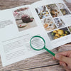 A hand holds a green magnifying glass over a page of an educational book featuring nature and food items.