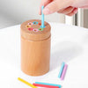 A hand is inserting a blue wooden rod into a sorting container filled with colorful wooden rings and colored rods.