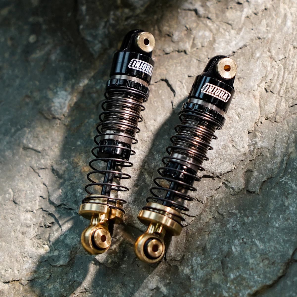 A pair of black and gold heavy-duty oil shocks for 1/24 RC crawlers resting on a gray rock surface.