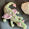 A soft green and pink rainbow dragon plush toy lying on a gray fabric surface.