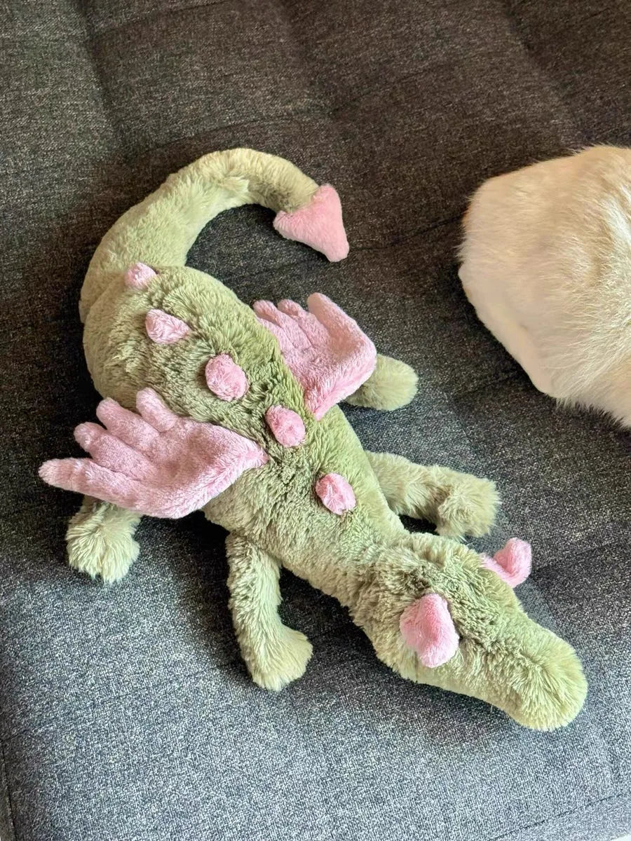 A soft green and pink rainbow dragon plush toy lying on a gray fabric surface.