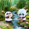 A cute blind box panda collectible figure with white and black coloring sitting near a small stream surrounded by green folia