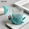 A teal handwoven crochet lizard coaster is displayed next to a glass and a skein of yarn on a white surface.