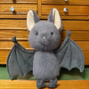 A soft grey bat plush toy with large ears and spread wings is displayed on a wooden surface.