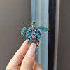 A hand holds a sparkling blue rhinestone turtle brooch pin against a light background.
