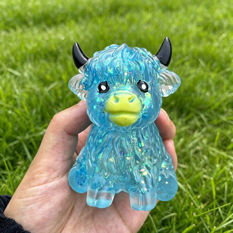 A hand holds a translucent blue glitter cow squishy stress relief fidget toy with horns in a grassy outdoor setting.