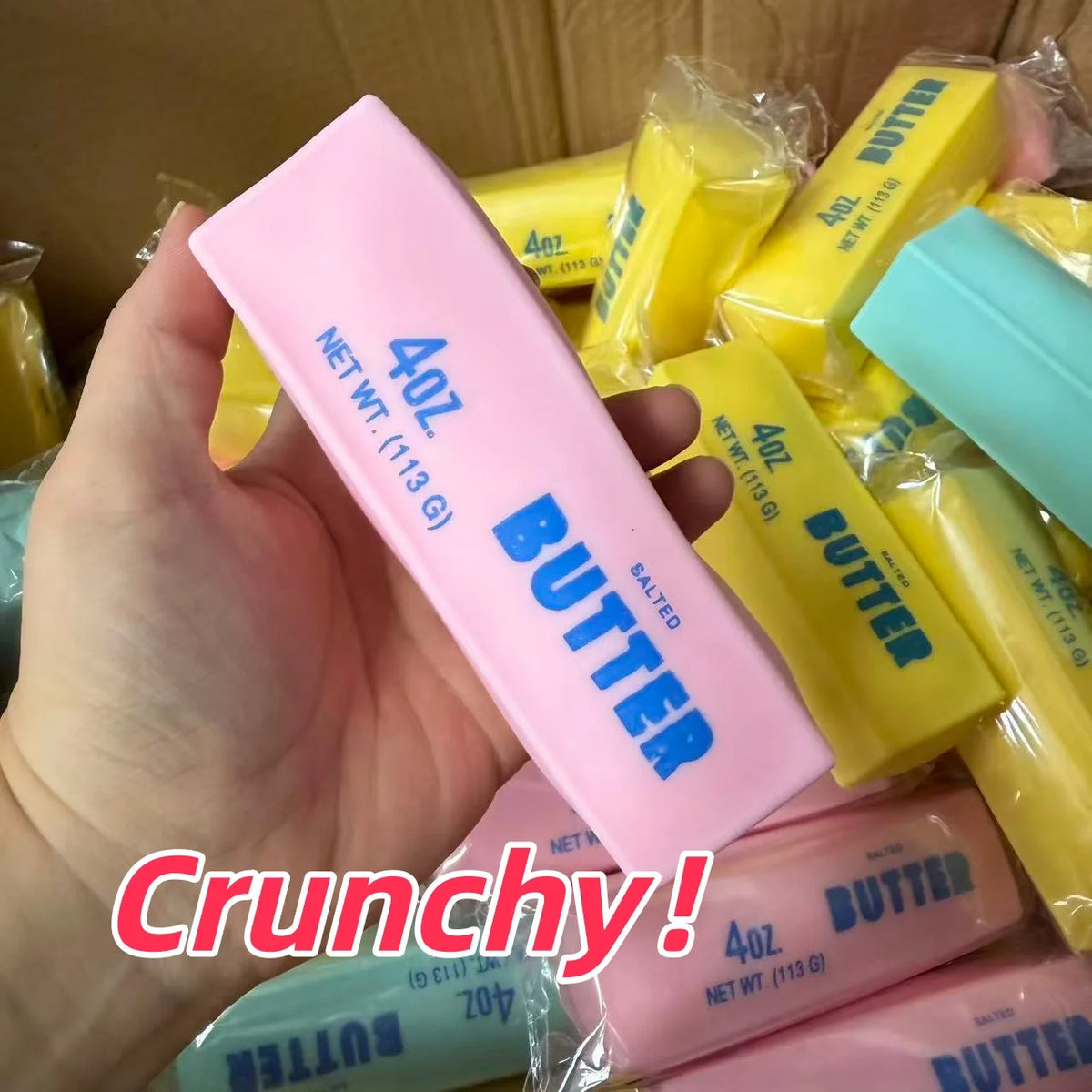 A hand holds a pink butter stick squishy stress ball next to several yellow butter sticks.
