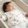 A baby is lying down in a white crib, resting on a white muslin washcloth with small animal designs.