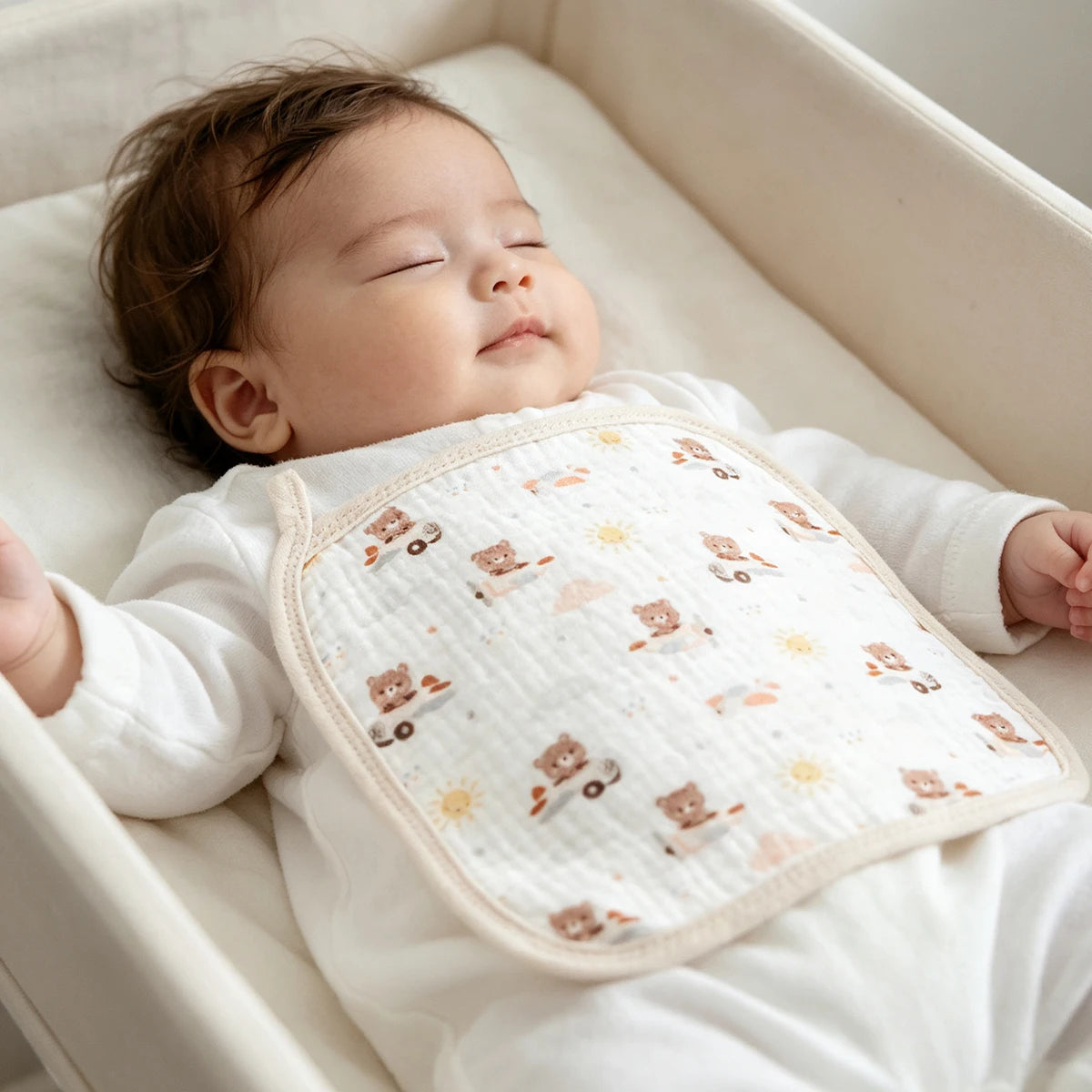 A baby is lying down in a white crib, resting on a white muslin washcloth with small animal designs.