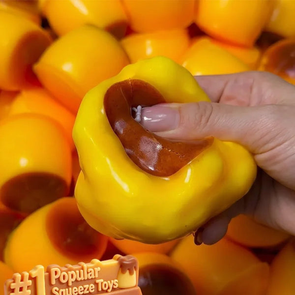 A hand holds a bright yellow silicone stress ball with an orange caramel-colored squeeze feature against a background of simi