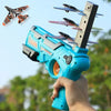 A hand holds a blue airplane launcher toy with three foam planes flying in the air against a bright sky.