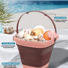 A pink expandable sand bucket toy with a comfort handle is shown outdoors on a beach setting.