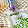 A purple diamond painting pen is applying green beads onto a white tray with small holes.