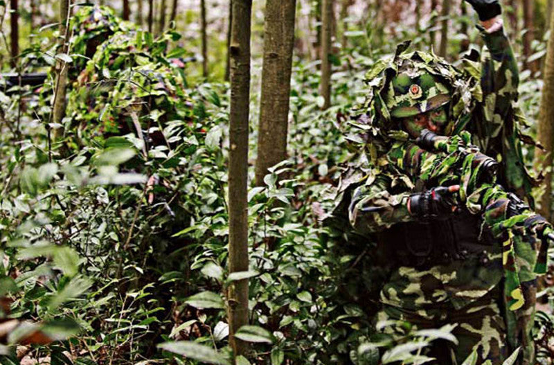 A person in a camouflage army costume set with accessories is partially visible amidst dense green foliage and tree trunks.