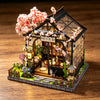 A miniature Japanese tea house diorama featuring a dark wooden structure, pink blossoms, and small plants.