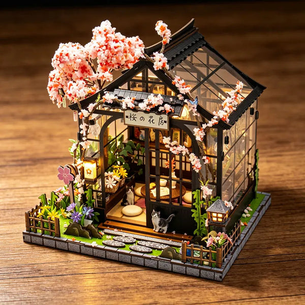 A miniature Japanese tea house diorama featuring a dark wooden structure, pink blossoms, and small plants.