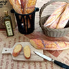 A miniature baguette is shown next to olive oil and a woven basket of bread in a rustic setting.
