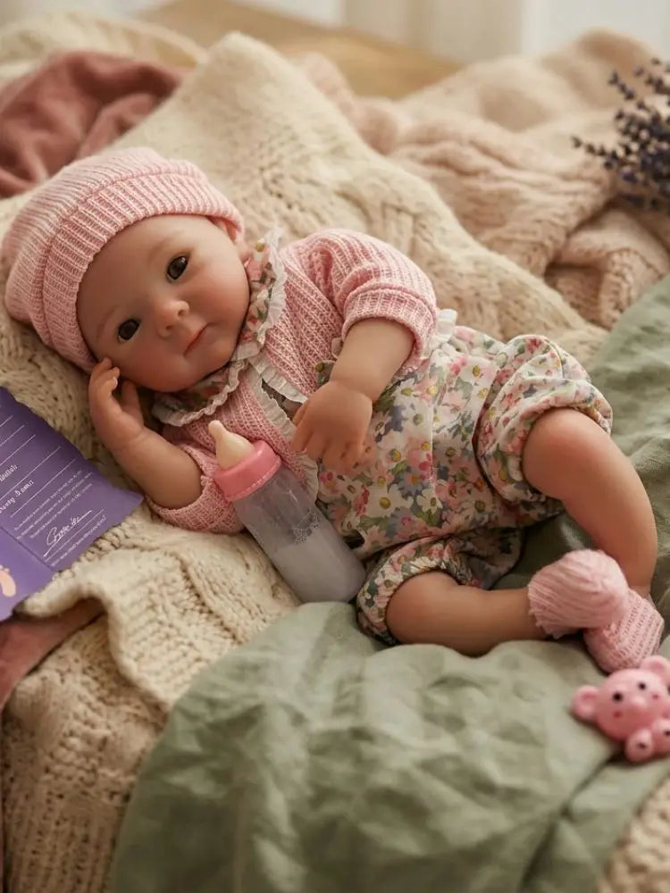 A realistic soft silicone reborn baby doll wearing a pink outfit, resting on a cream-colored knitted blanket.