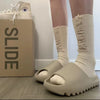 A pair of beige EVA platform slides with fluffy white cuff socks are displayed next to a cardboard box.