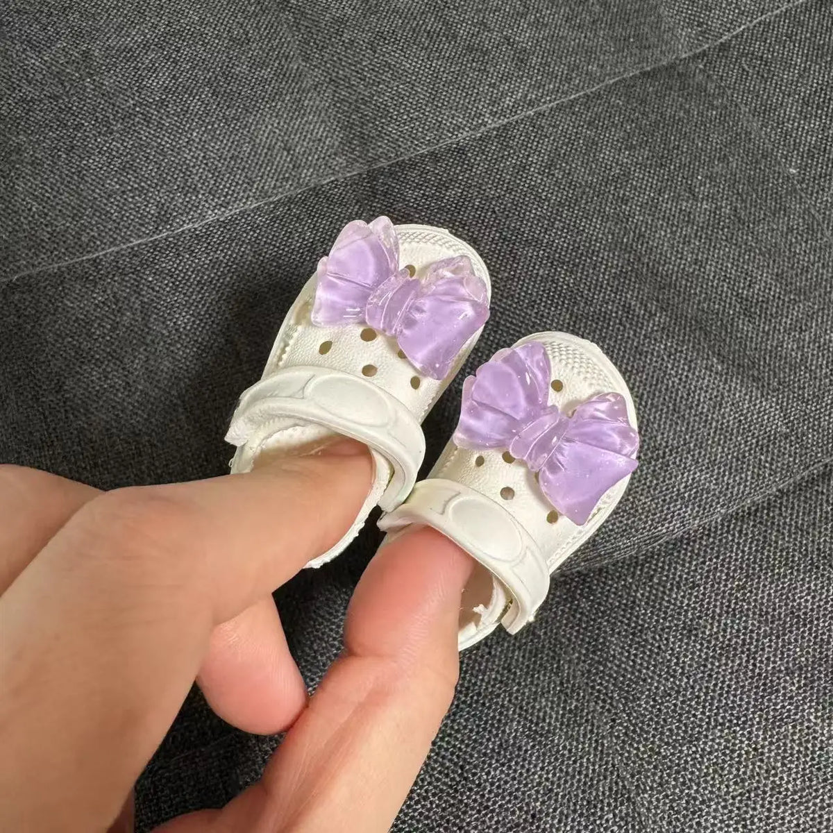 A pair of white fashion slippers adorned with purple bows being held by a hand.