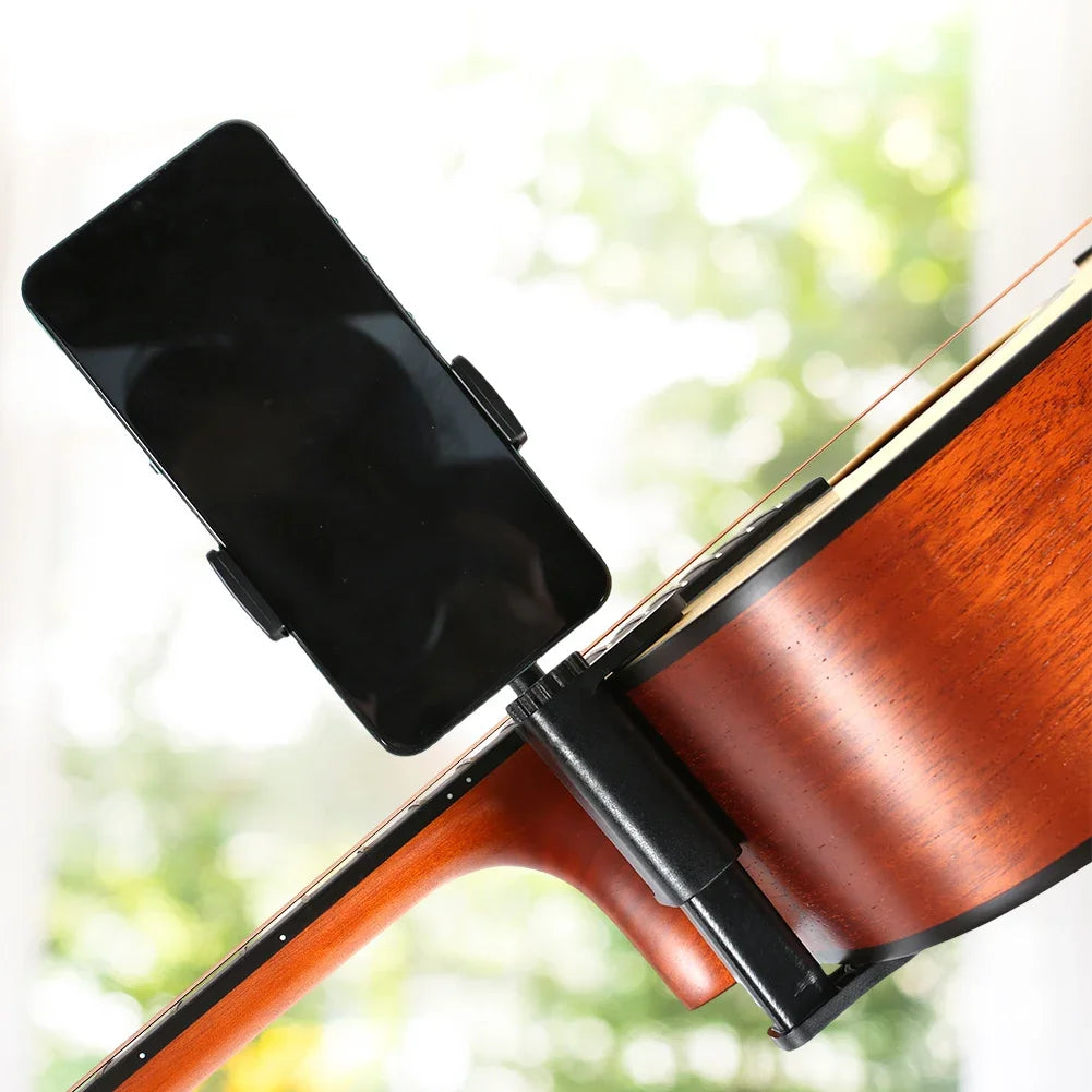 A black smartphone is mounted on a clamp attached to the neck of a wooden guitar.