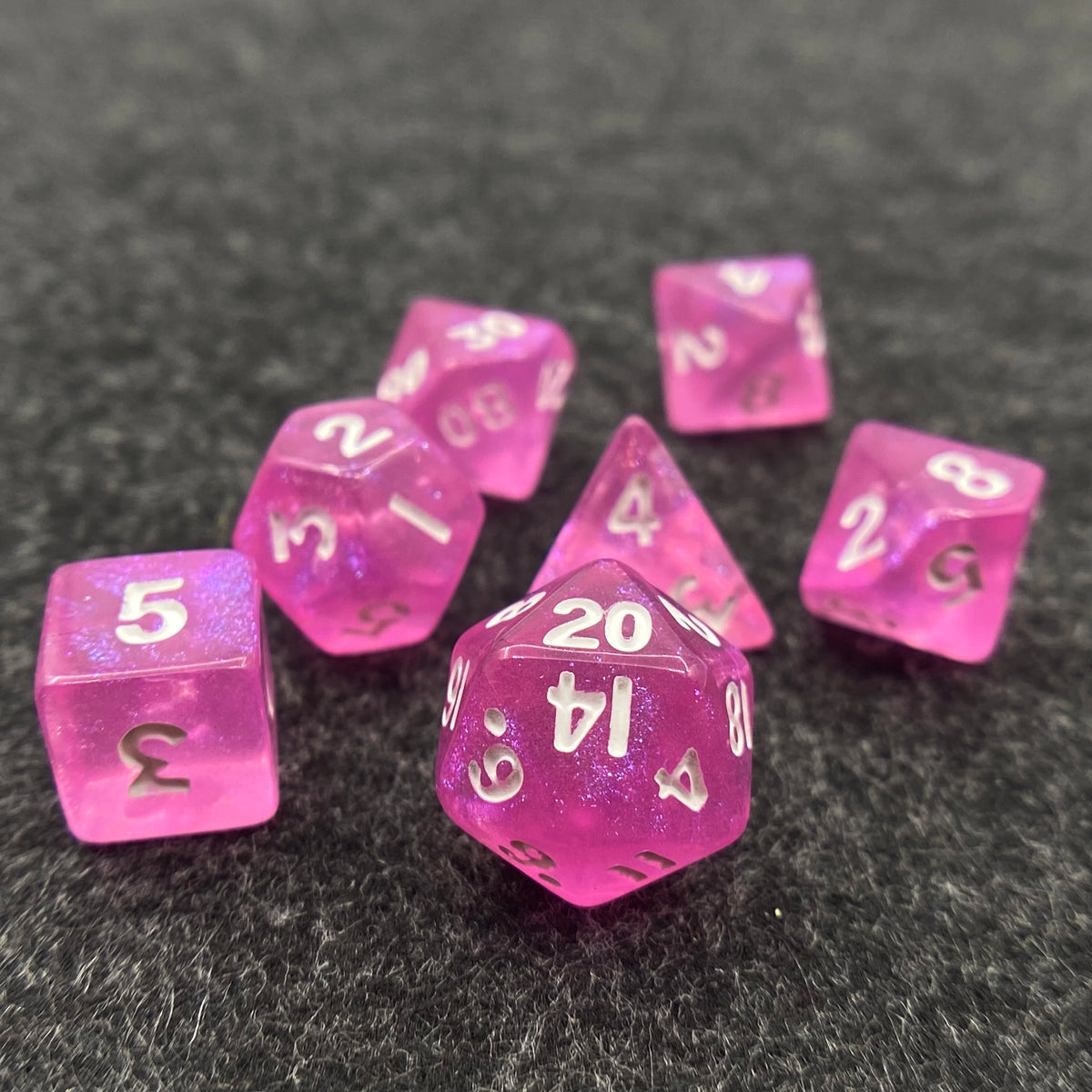 A collection of seven iridescent pink acrylic polyhedral dice scattered on a dark surface.