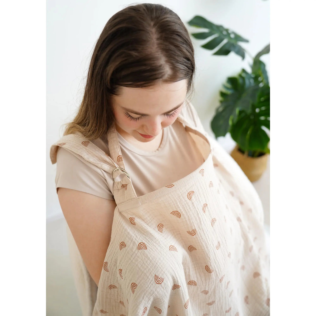 A woman wearing a beige breathable muslin nursing cover with a delicate pattern, looking down.