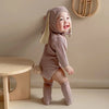 A baby wearing a light pink knitted rabbit romper set with a matching hat and socks standing in a studio setting.