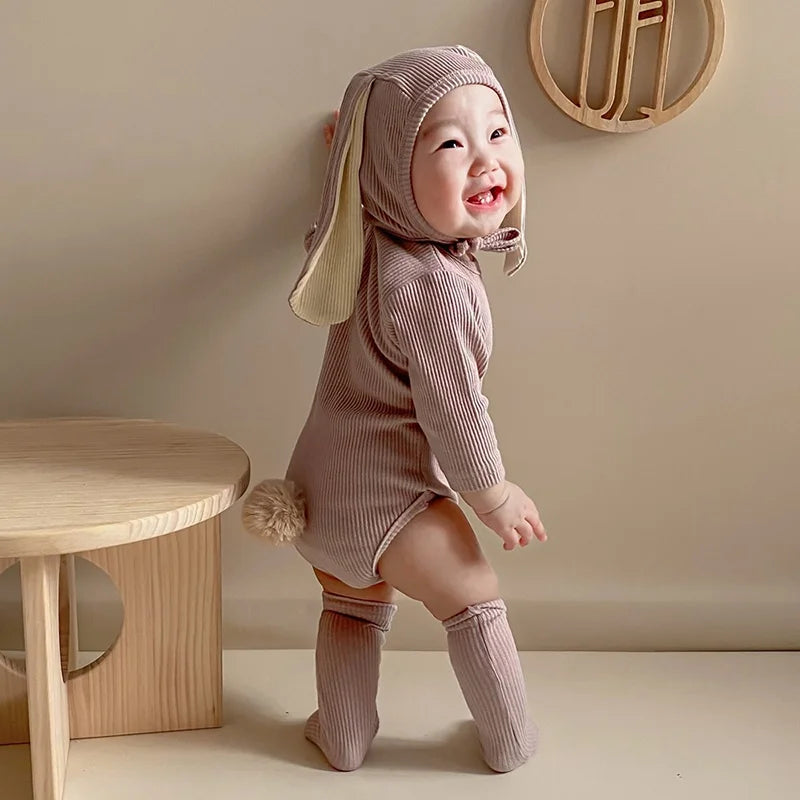 A baby wearing a light pink knitted rabbit romper set with a matching hat and socks standing in a studio setting.