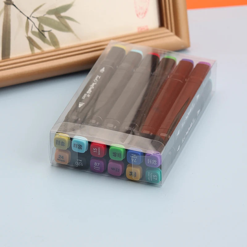 A clear plastic box of dual-tip alcohol art markers with various colors and tips displayed on a light blue surface.