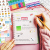 A hand holds a pink notebook with various planner stickers and a pen, next to a colorful grid of planner stickers.