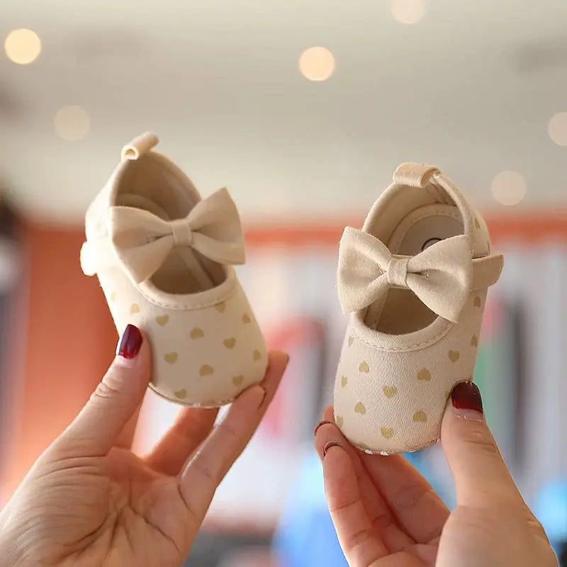A pair of beige soft sole first walker shoes with a bow detail being held by a person.