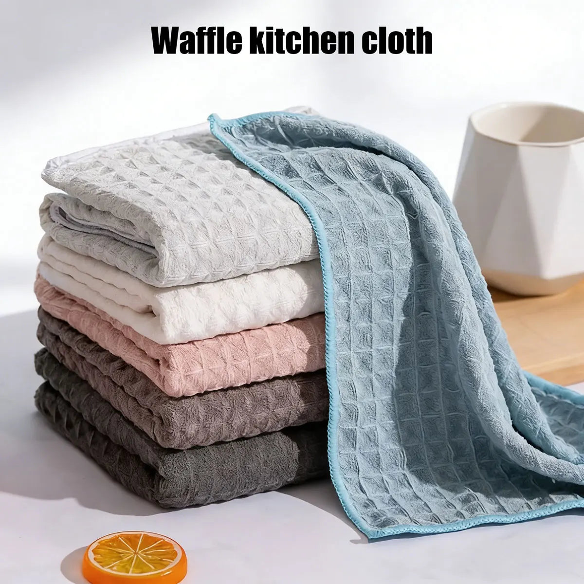A stack of light-colored and gray waffle kitchen cloths, including a blue-green cloth, next to an orange slice.