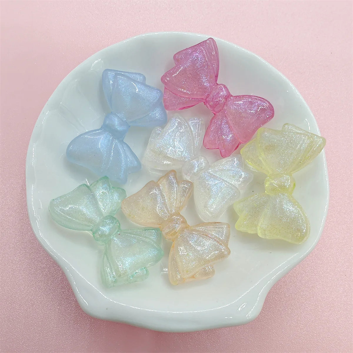 A collection of shimmering pink, blue, and yellow mermaid bowknot beads for jewelry making displayed on a white dish.