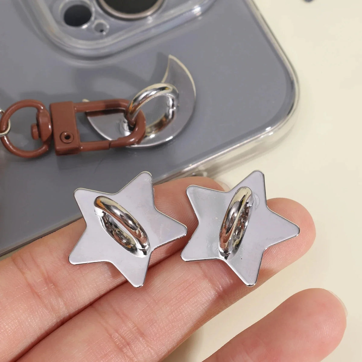 Silver star-shaped phone charm hook with an adhesive backing being held by a hand.