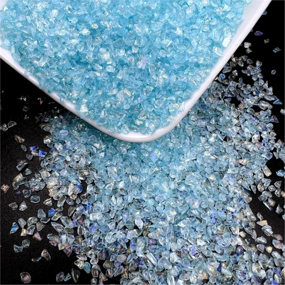 A pile of light blue crushed glass crystals in a white dish and scattered on a dark surface.
