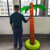 A person is holding an oversized inflatable palm tree with green fronds and an orange trunk in a brightly lit room.