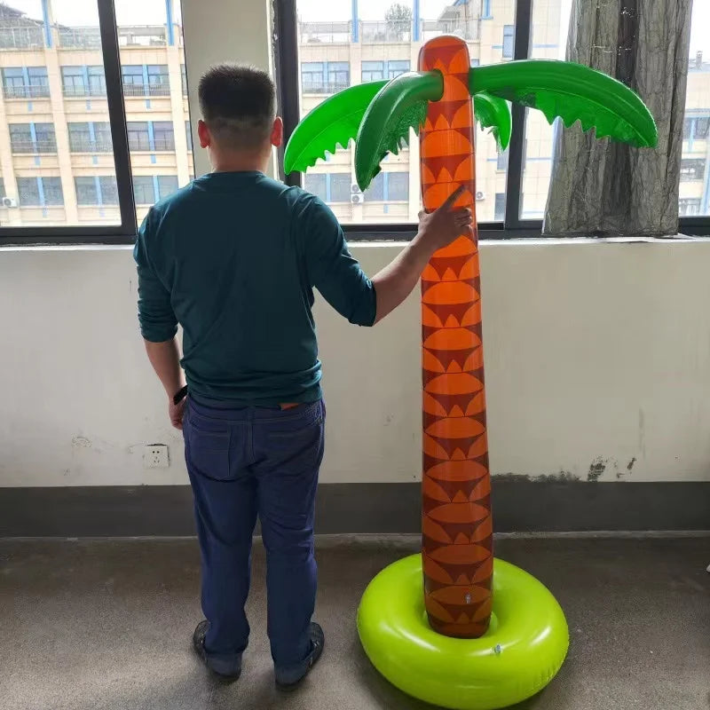 A person is holding an oversized inflatable palm tree with green fronds and an orange trunk in a brightly lit room.