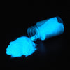 A small glass bottle of luminous glow-in-the-dark pigment powder next to a pile of the same bright blue powder against a blac